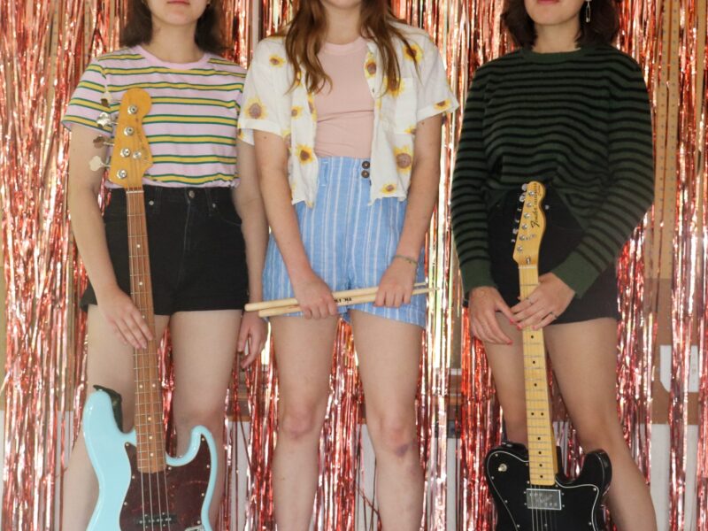 The three band members of UN-T.I.L. stand in front of a sparkly, pink, fringe background. From left to right: Lena Seavey, Tierney Coughlin and Inara Seveay.