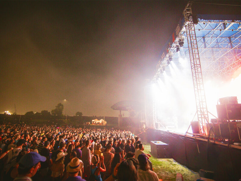 Dozens of fans are pressed together in front of a brightly lit outdoor stage