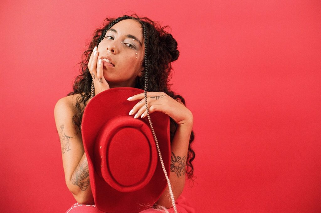 Indigo De Souza poses in front of a red background, holding a red cowboy hat.